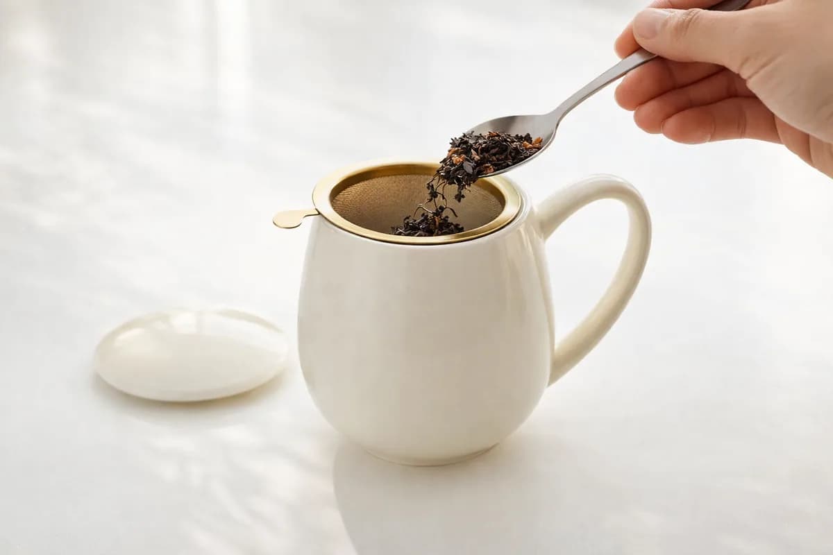 Dodavanje čaja u zlatni filter iznad bijele šalice.